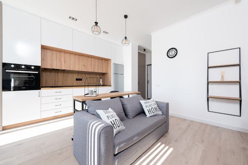 Interior of bright modern room with open kitchen with cupboards and oven next to couch with pillows and table