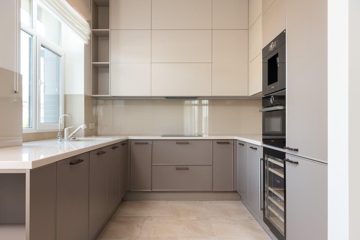 Interior of bright modern kitchen with cupboards and oven with microwave near sink with tap next to window