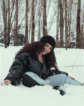 Woman in winter attire smiling while sitting in snow amidst trees.