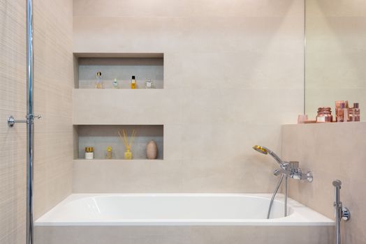 Interior of light modern bathroom with white bath with shower under shelves with decorative objects next to mirror