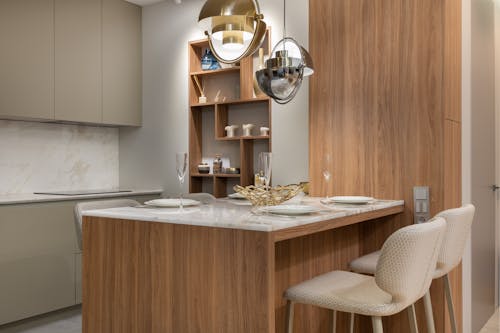 Interior of bright modern kitchen with cupboards near chairs with counter decorated with plates and glasses under lamps next to shelves