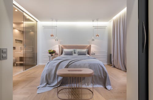 Interior of modern light home with bed with pillows and blanket next to table under lamps near pouf and glass door to washroom