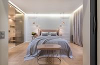 Interior of modern light home with bed with pillows and blanket next to table under lamps near pouf and glass door to washroom