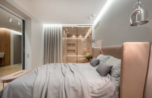 Interior of modern light bedroom with pillows and blanket on bed under lamps next to pouf near glass wall with curtain