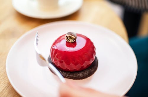 High angle of crop anonymous person eating delicious cake covered with red mirror glaze and served on white plate during coffee break in cafe