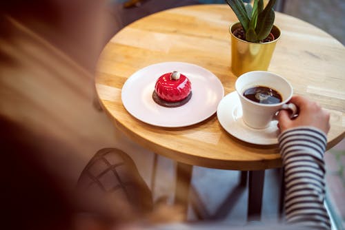 Free From above of crop anonymous lady in casual outfit drinking aromatic coffee and eating yummy glaze cake while sitting at round wooden table in modern cafe Stock Photo