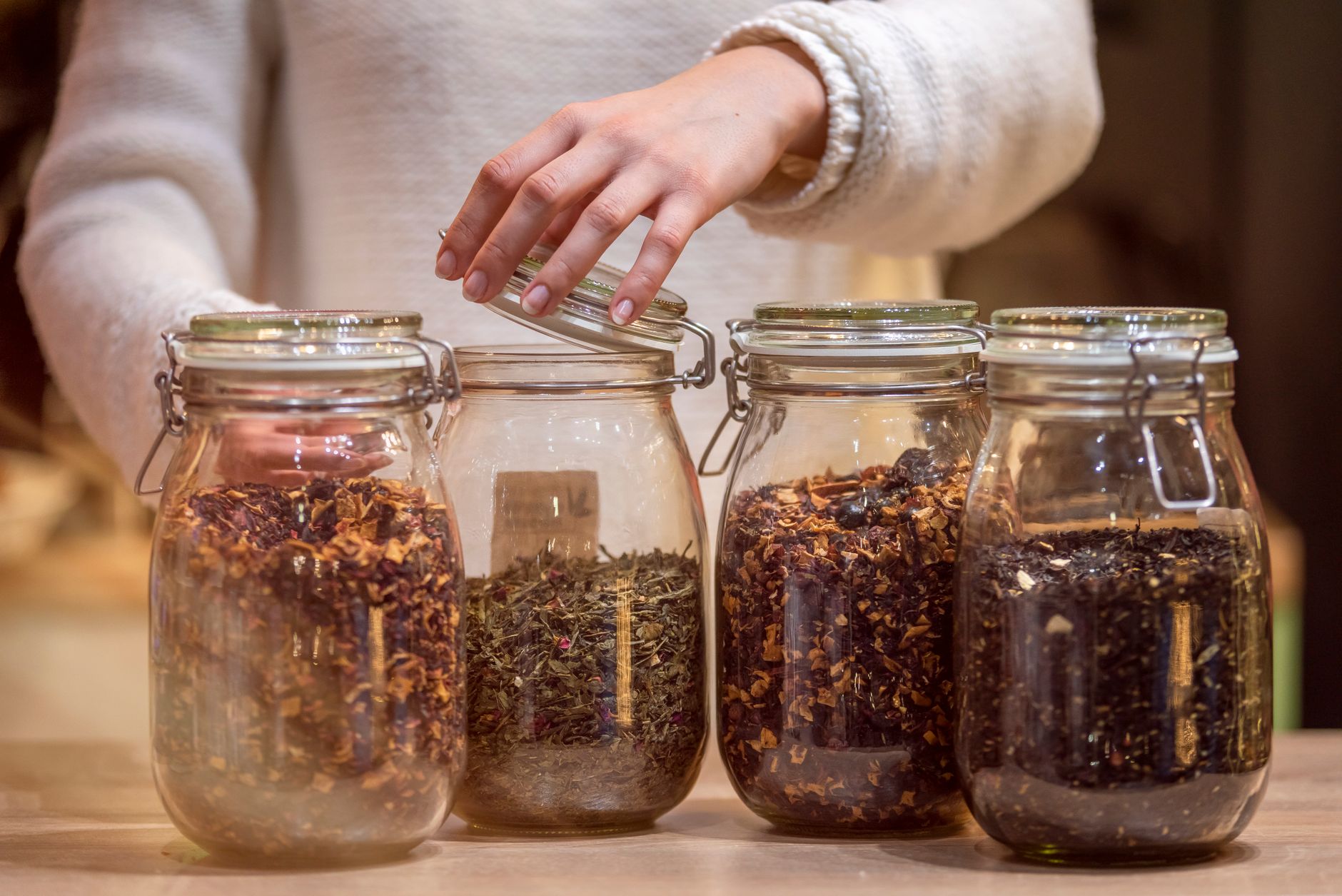 Crop unrecognizable woman in sweater standing at wooden table and opening jar with aromatic tea against blurred backgrou