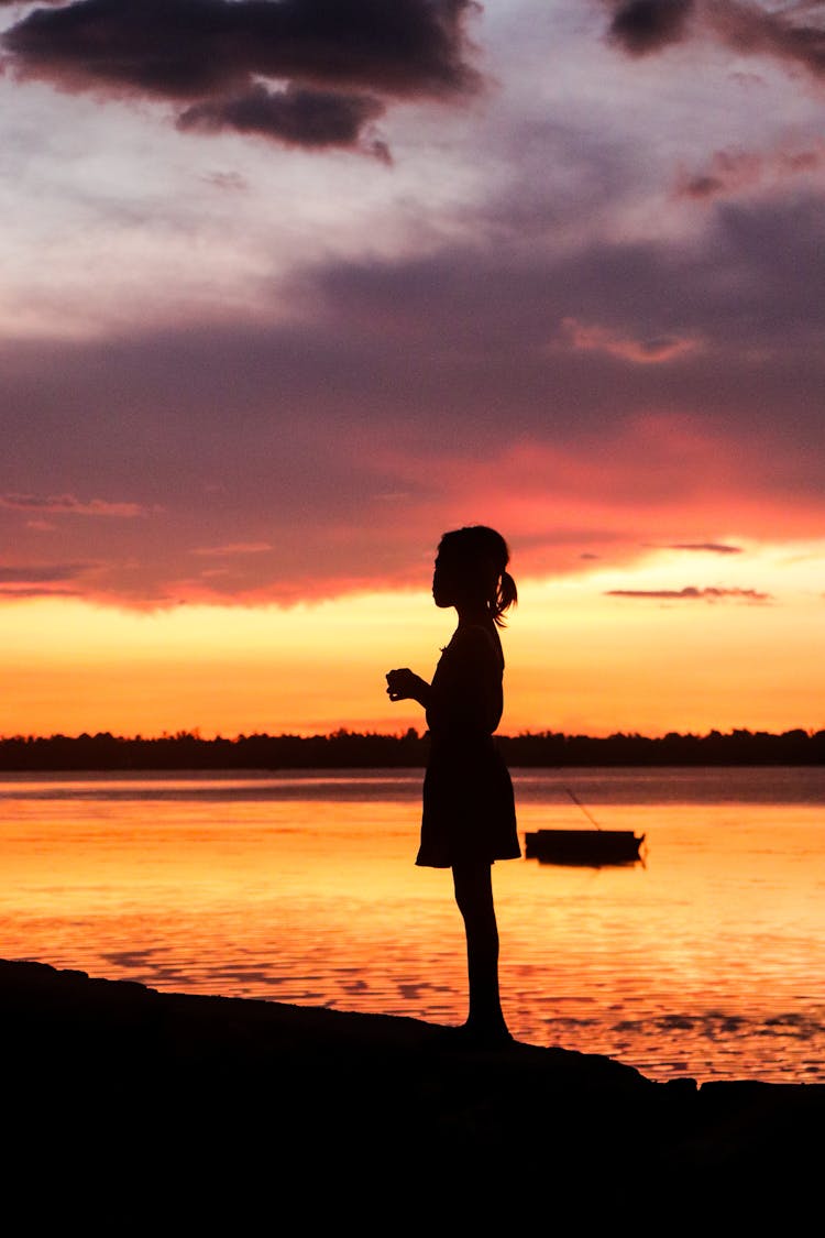 Silhouette Of Little Girl On Lake Shore At Sunset