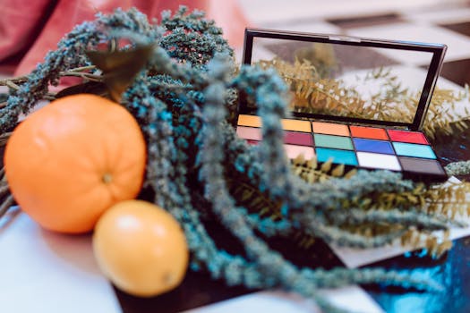 A colorful makeup palette amidst citrus fruits and green foliage, creating a vibrant artistic scene.