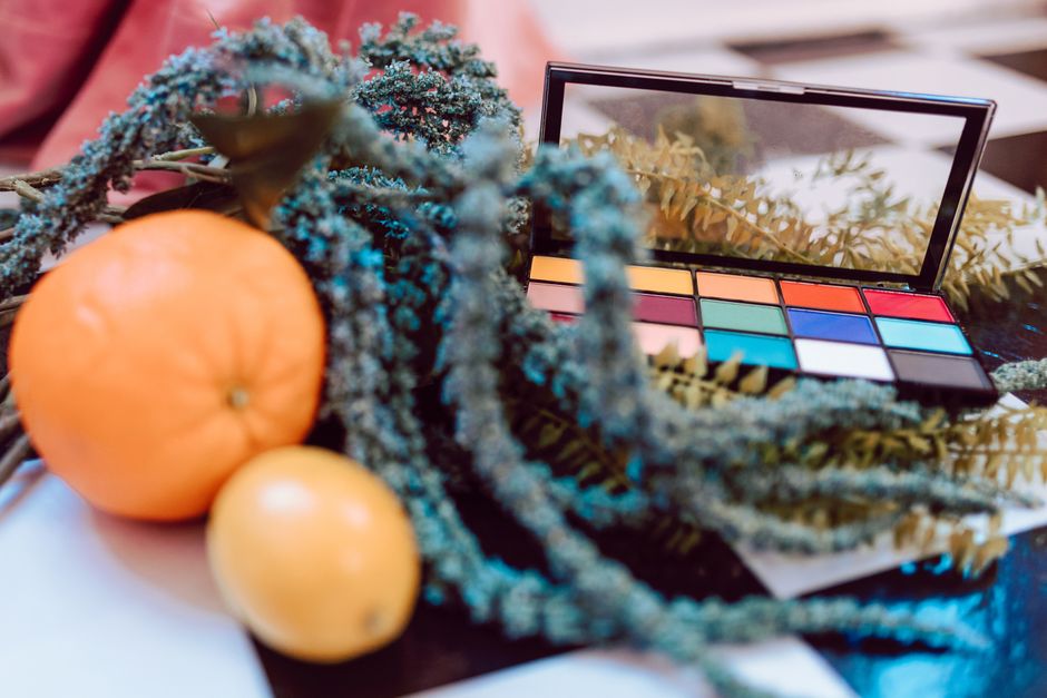 A colorful makeup palette amidst citrus fruits and green foliage, creating a vibrant artistic scene.