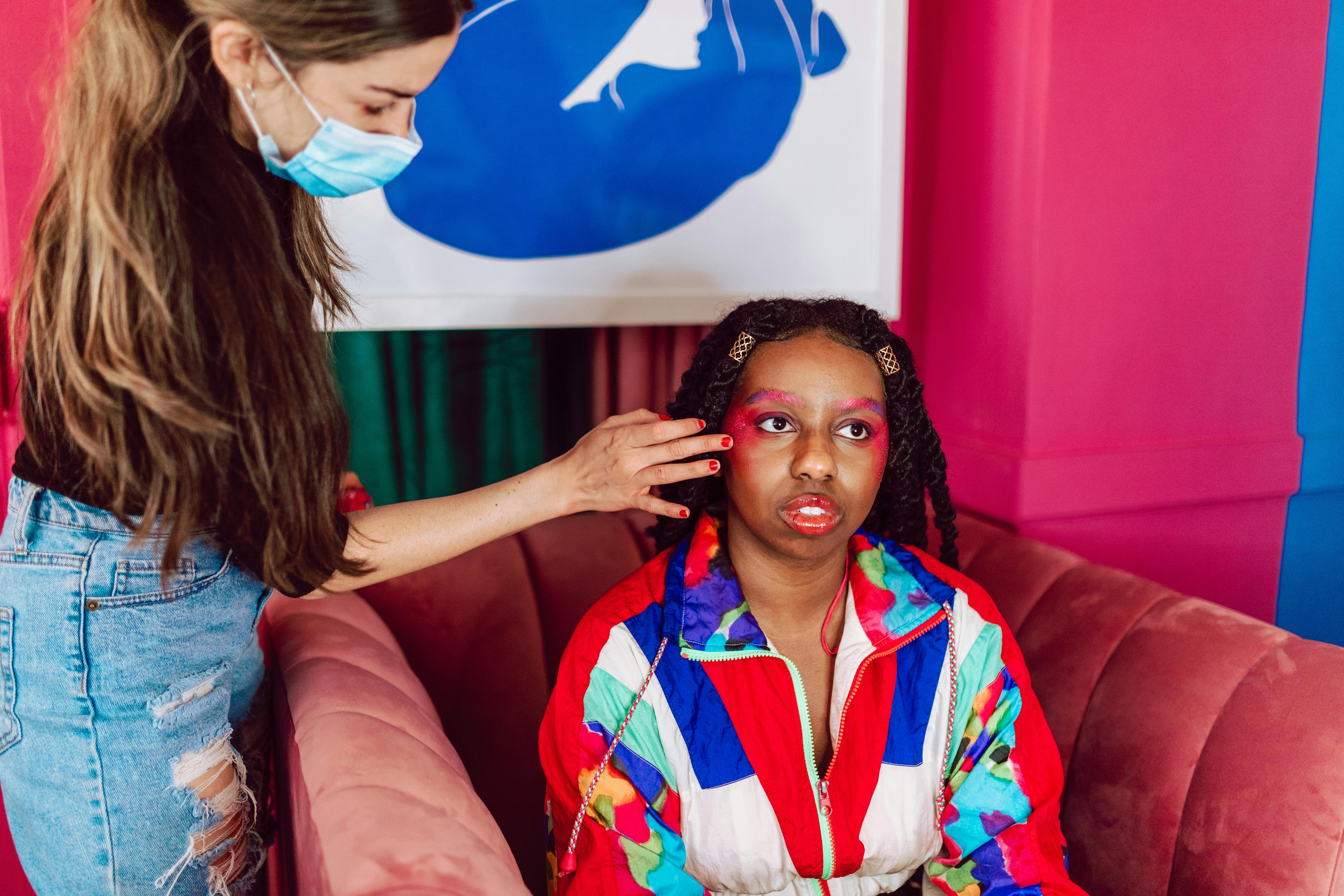 Makeup artist wearing a mask applies makeup to a young woman on a couch in a colorful interior.