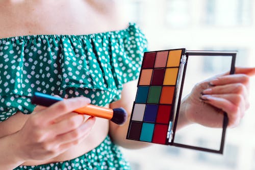 Person in Green Tube Top Holding an Eyeshadow Palette and Makeup Brush