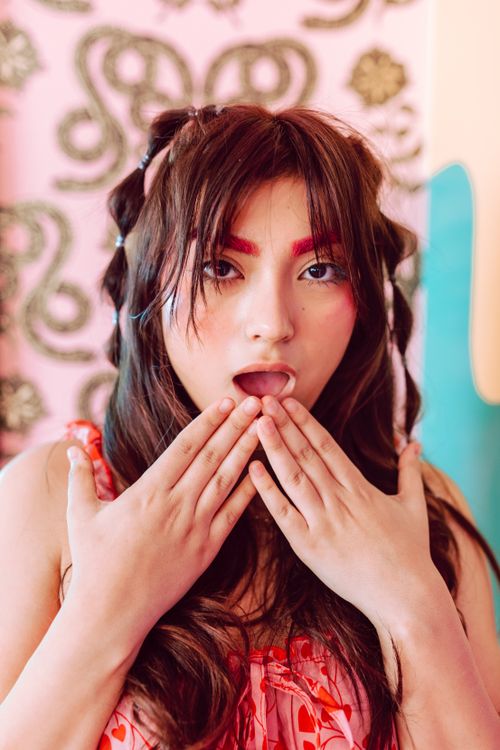 Free A young woman with vibrant makeup poses playfully indoors with hands on face. Stock Photo