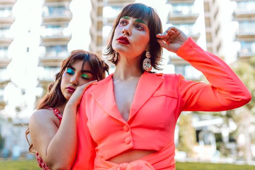 Two fashionable women with bold makeup pose outdoors in stylish outfits.