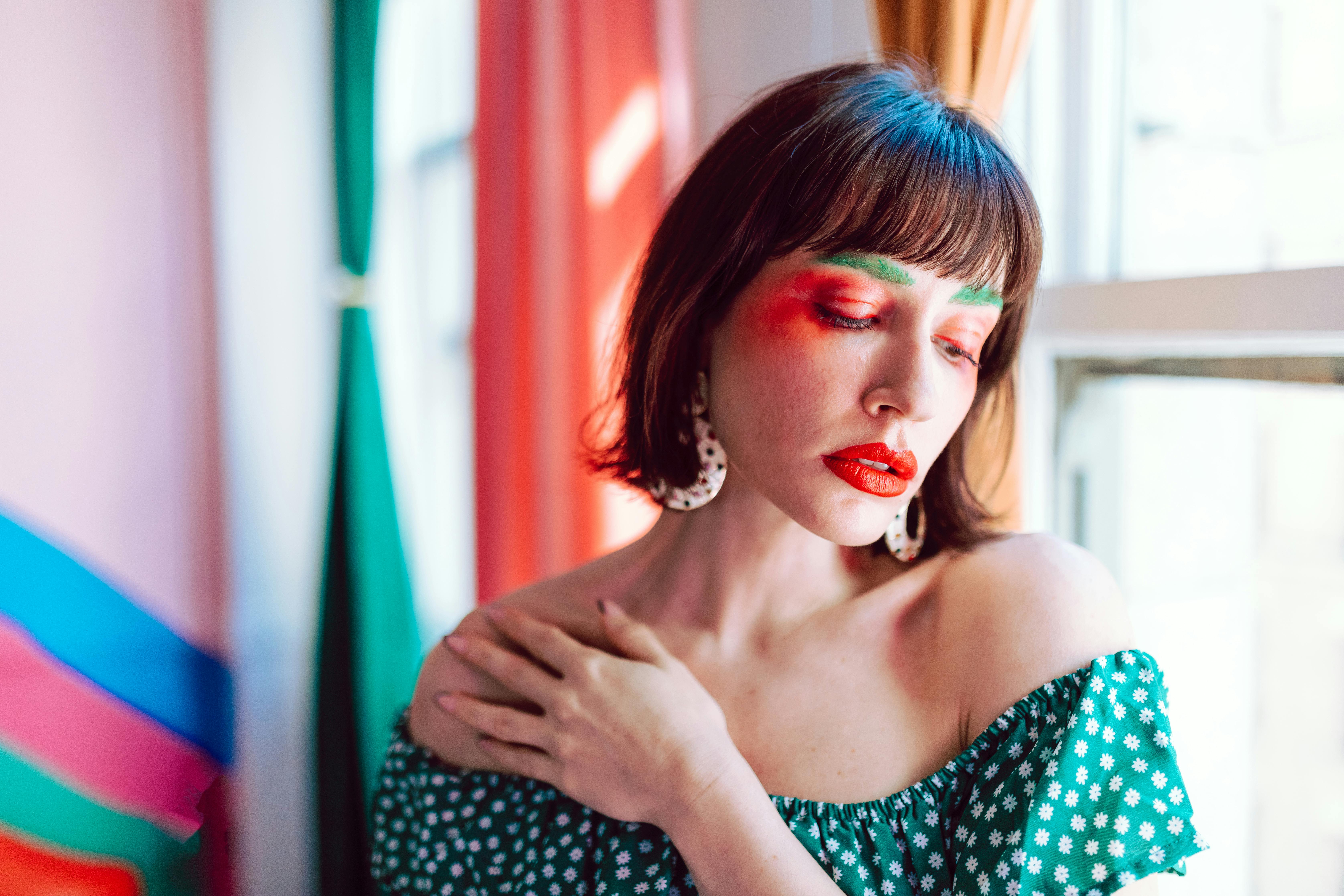 Colorful portrait of a woman in floral off-shoulder top with vibrant makeup near window.