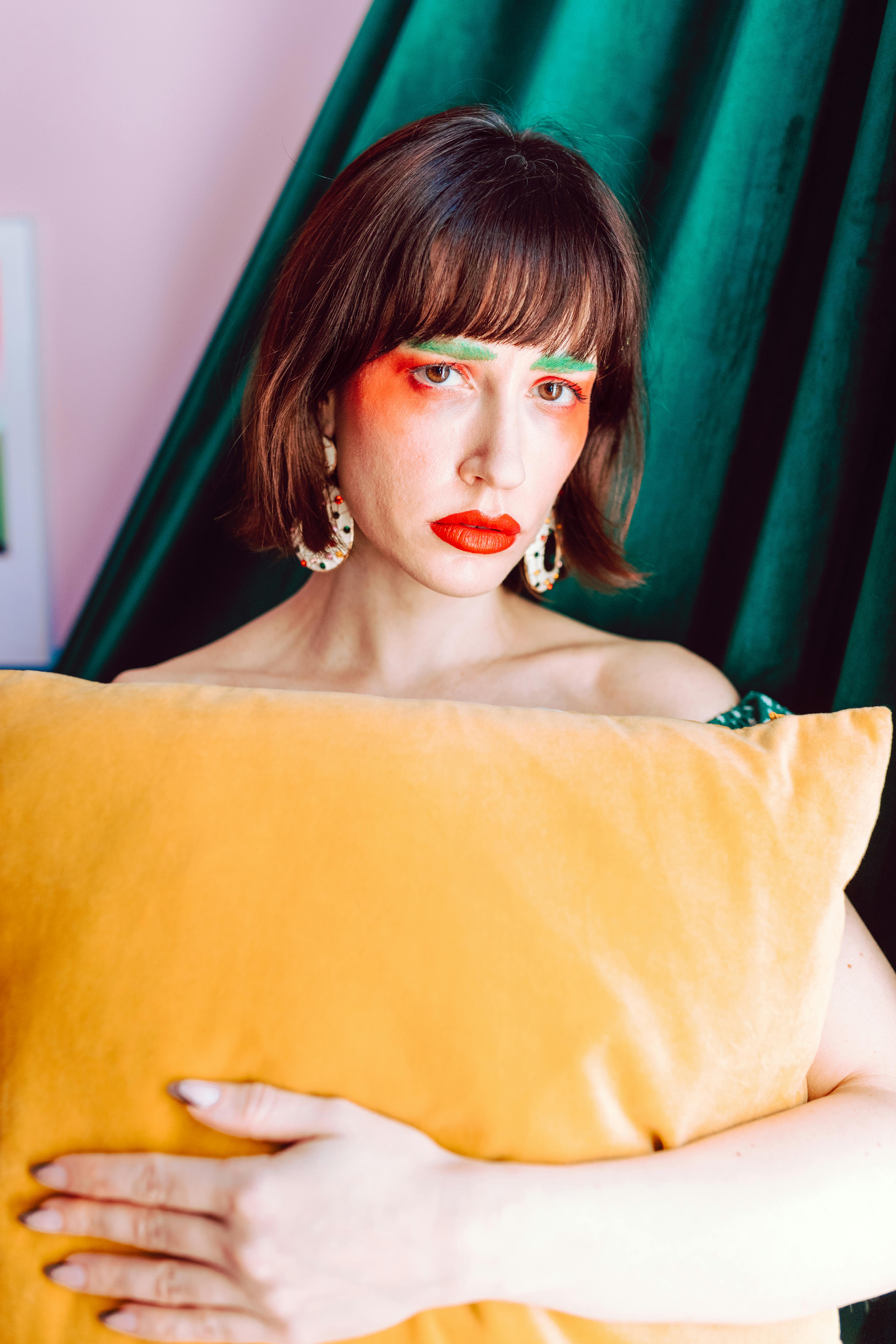 Young adult with vibrant makeup holding a yellow pillow against colorful background.