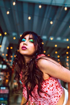 A fashionable woman with vibrant makeup poses under decorative string lights at night.