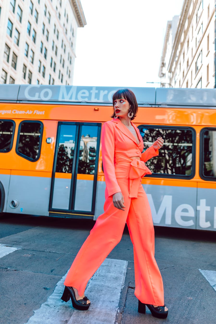 Stylish woman in orange suit confidently striding across city crosswalk with metro bus background.