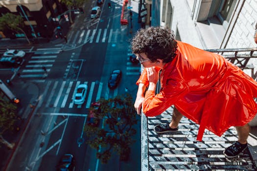 Man in vibrant red coat overlooks city from tall building, showcasing bold urban fashion.
