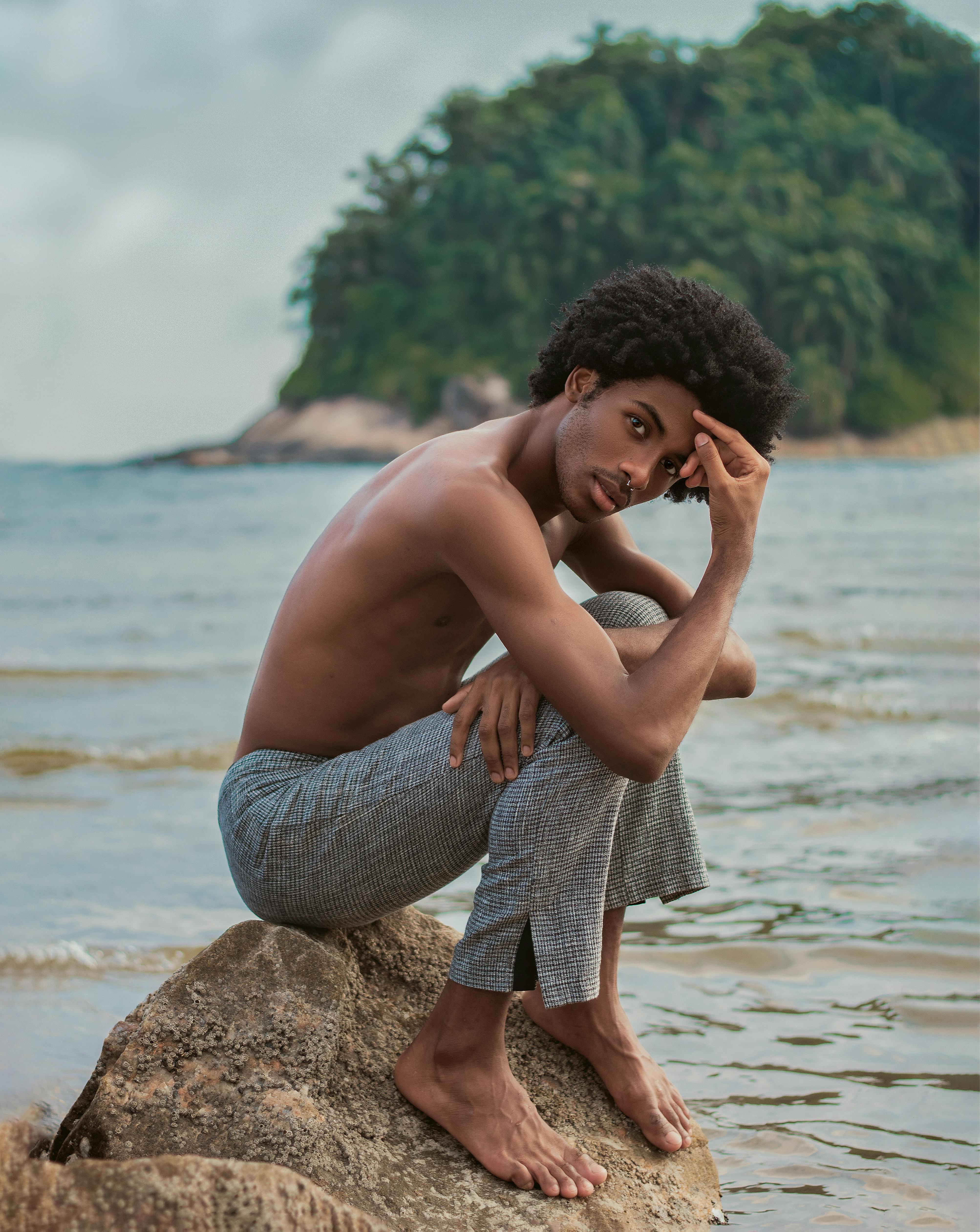 Side view full body of serious African American shirtless barefoot male with naked torso sitting on stone near waving river and looking at camera