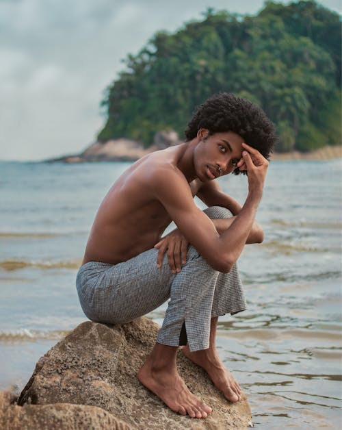 Side view full body of serious African American shirtless barefoot male with naked torso sitting on stone near waving river and looking at camera