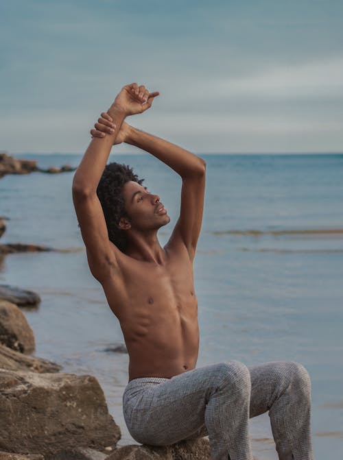 Shirtless peaceful African American male with hands above head sitting on stone near endless sea and enjoying freedom