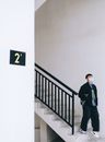 Full body of concentrated male in medical mask and casual clothes with hand in pockets getting down white staircase with contrast black railings