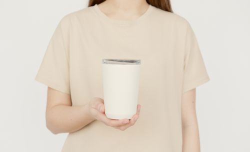 Free Young adult in a beige t-shirt holding a blank white tumbler against a neutral background. Stock Photo