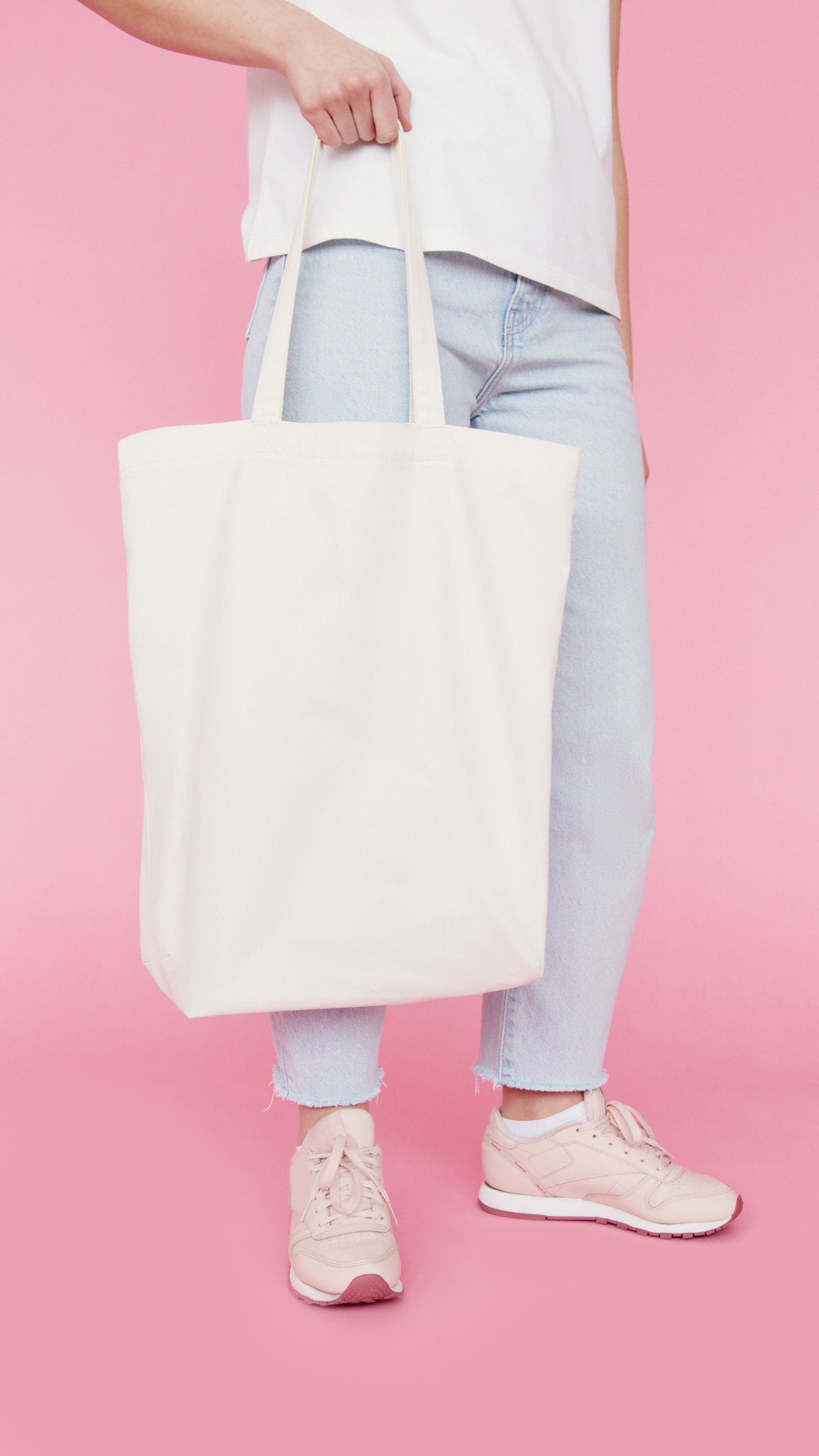 Jovem mulher elegante segurando uma bolsa de lona branca contra um fundo rosa pastel.