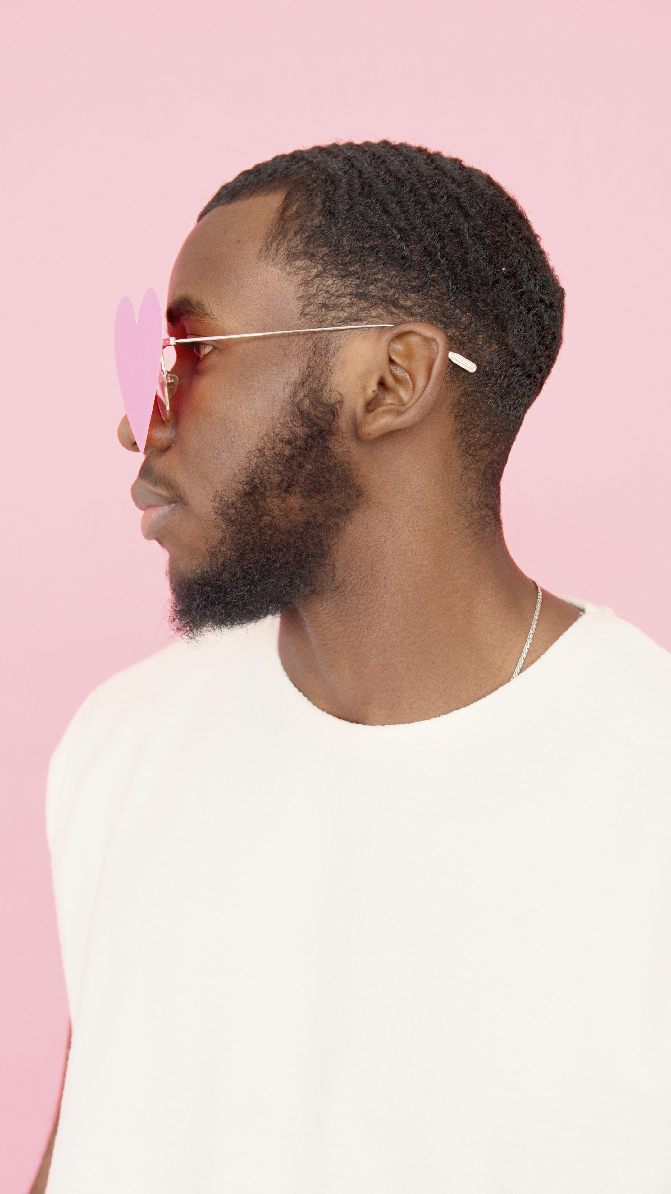 A fashionable man in a white t-shirt with heart-shaped sunglasses on a pink backdrop.