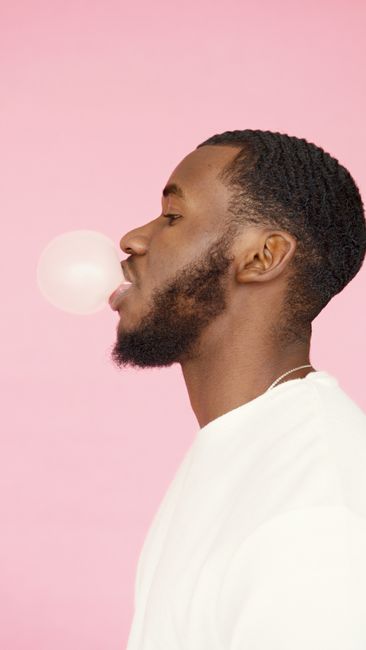 Side profile of a man with a beard blowing bubblegum on a pastel pink background.