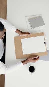 Top view of person arranging empty paper on clipboard with coffee and books nearby.