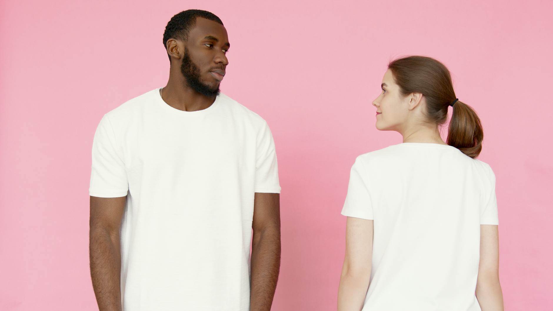 A man and woman in white t-shirts stand against a pink backdrop.