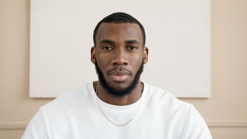 A composed Black man in his 30s with a short beard, wearing a white T-shirt and a thin gold chain, looks directly at the camera against a plain neutral backdrop.