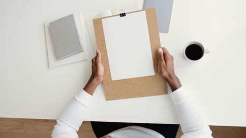 Free Top view of a person holding a clipboard with blank paper on a desk with coffee and books. Stock Photo