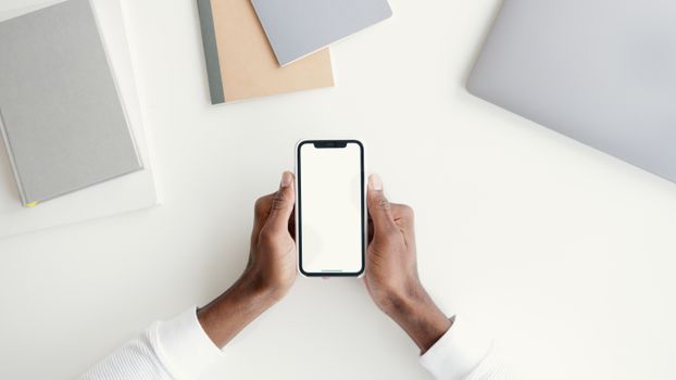 Hands holding smartphone with blank screen, surrounded by notebooks and a laptop on a white desk.
