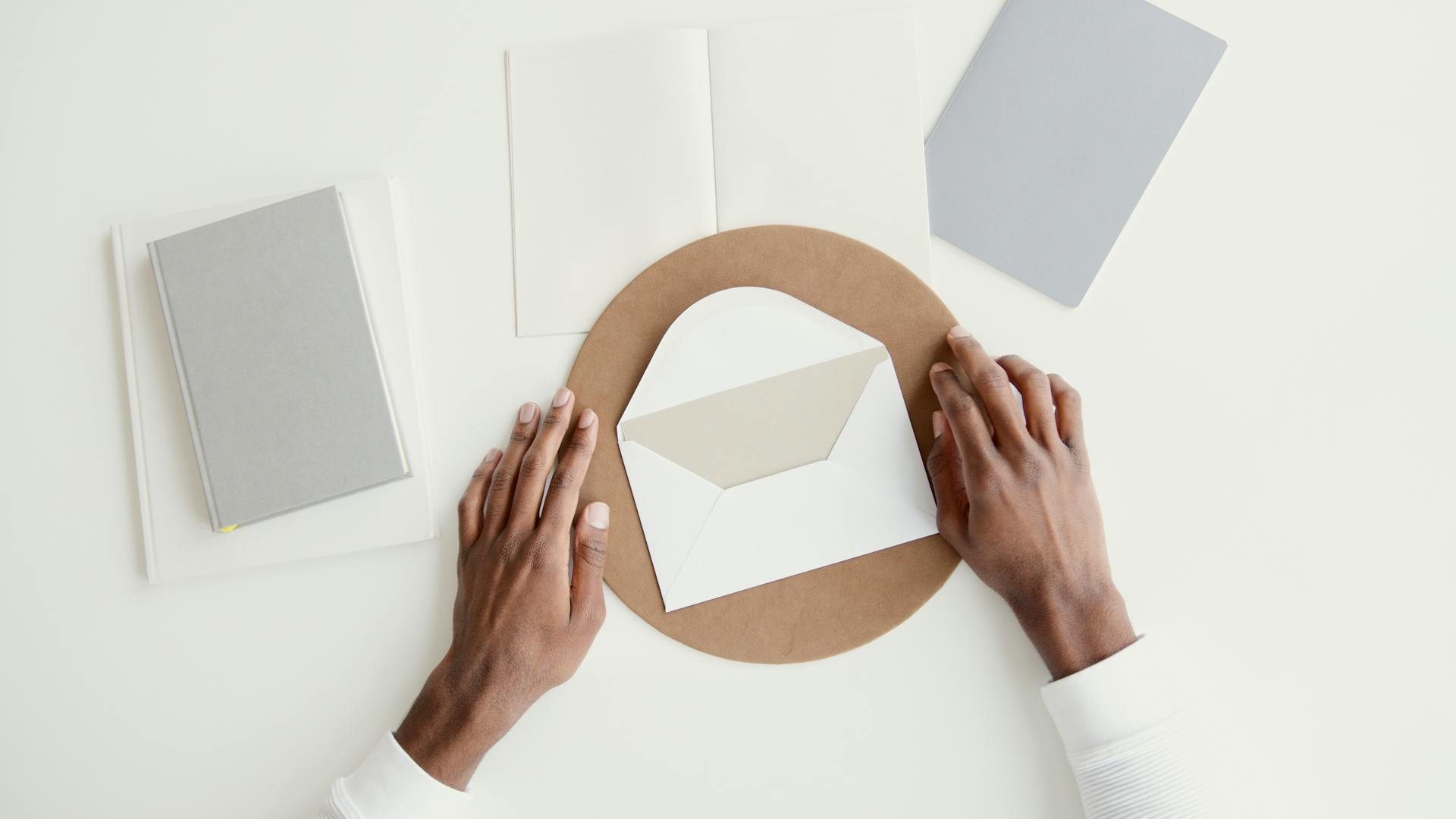 Overhead shot of hands arranging an envelope among notebooks on a minimal workspace.