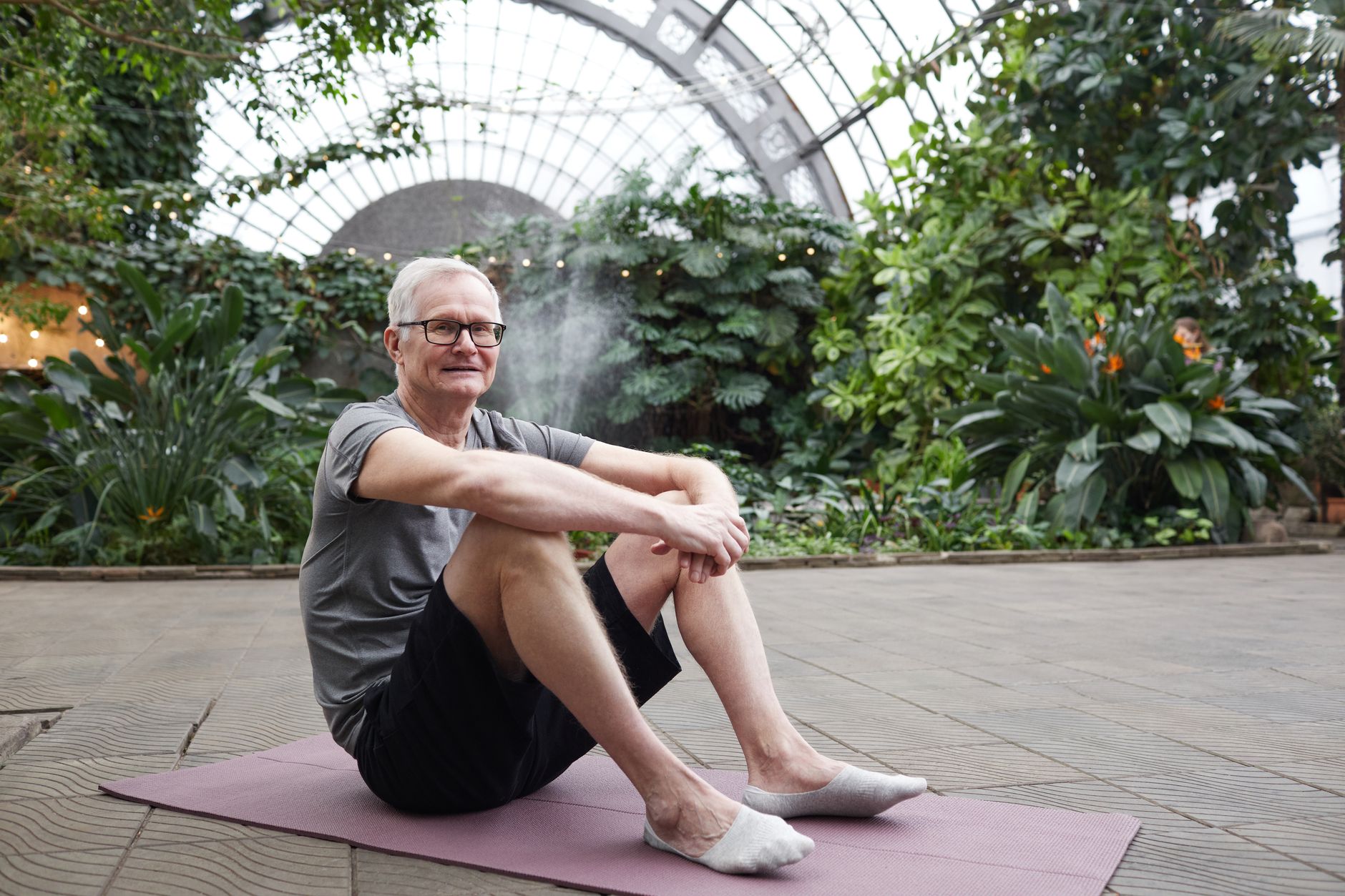 Older man enjoying yoga session on a mat in a lush botanical garden.