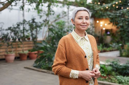 Free A gracefully smiling senior woman in a stylish cardigan poses in a serene indoor greenhouse. Stock Photo