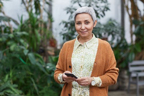 Free Elderly woman with smartphone standing in a greenhouse, smiling. Stock Photo