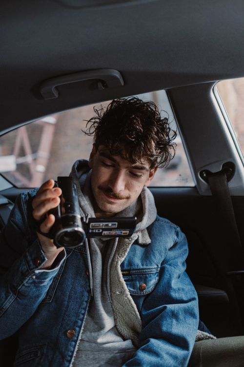 Free Young man with curly hair filming with a camcorder inside a car. Stock Photo
