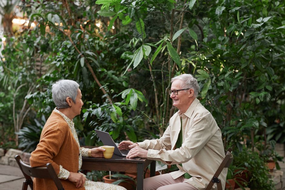 Happy senior couple sitting outdoors with laptop and coffee, enjoying nature.