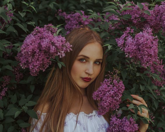 Portrait of peaceful young female with long straight hair wearing white dress touching blossoming flowers of lilac bush and looking at camera