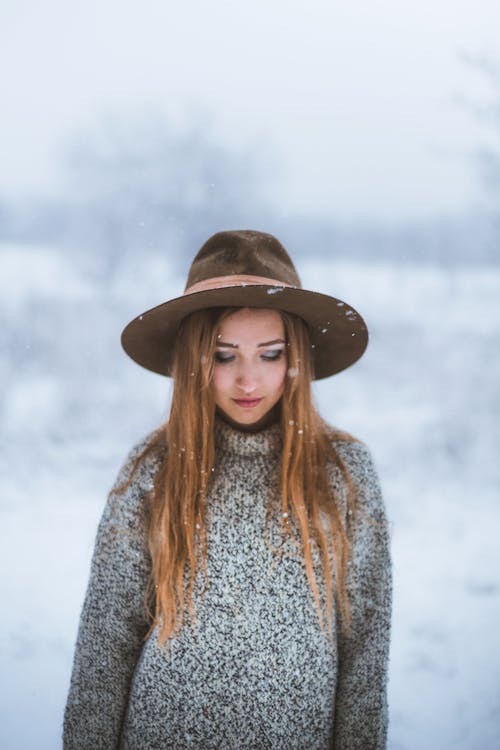 Young female with long straight hair in warm sweater and brown hat looking down while standing under snowfall in winter field