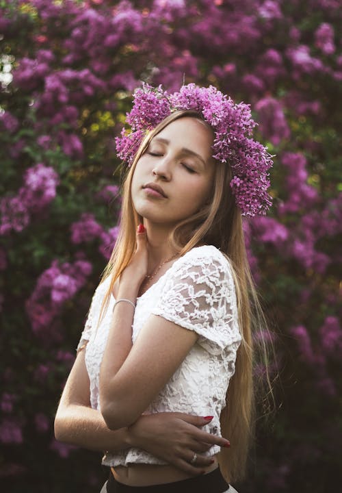 Calm gentle young lady in white lace blouse touching neck and enjoying smell of blooming lilac flowers