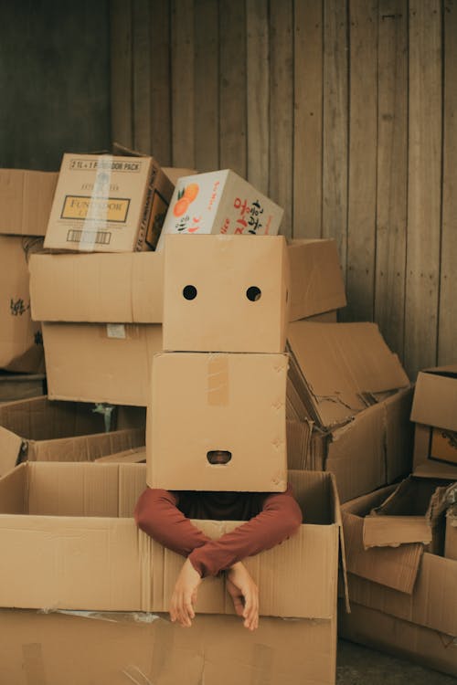 Free Faceless person sitting in big box with carton box on head wearing casual clothes near wall in light place Stock Photo
