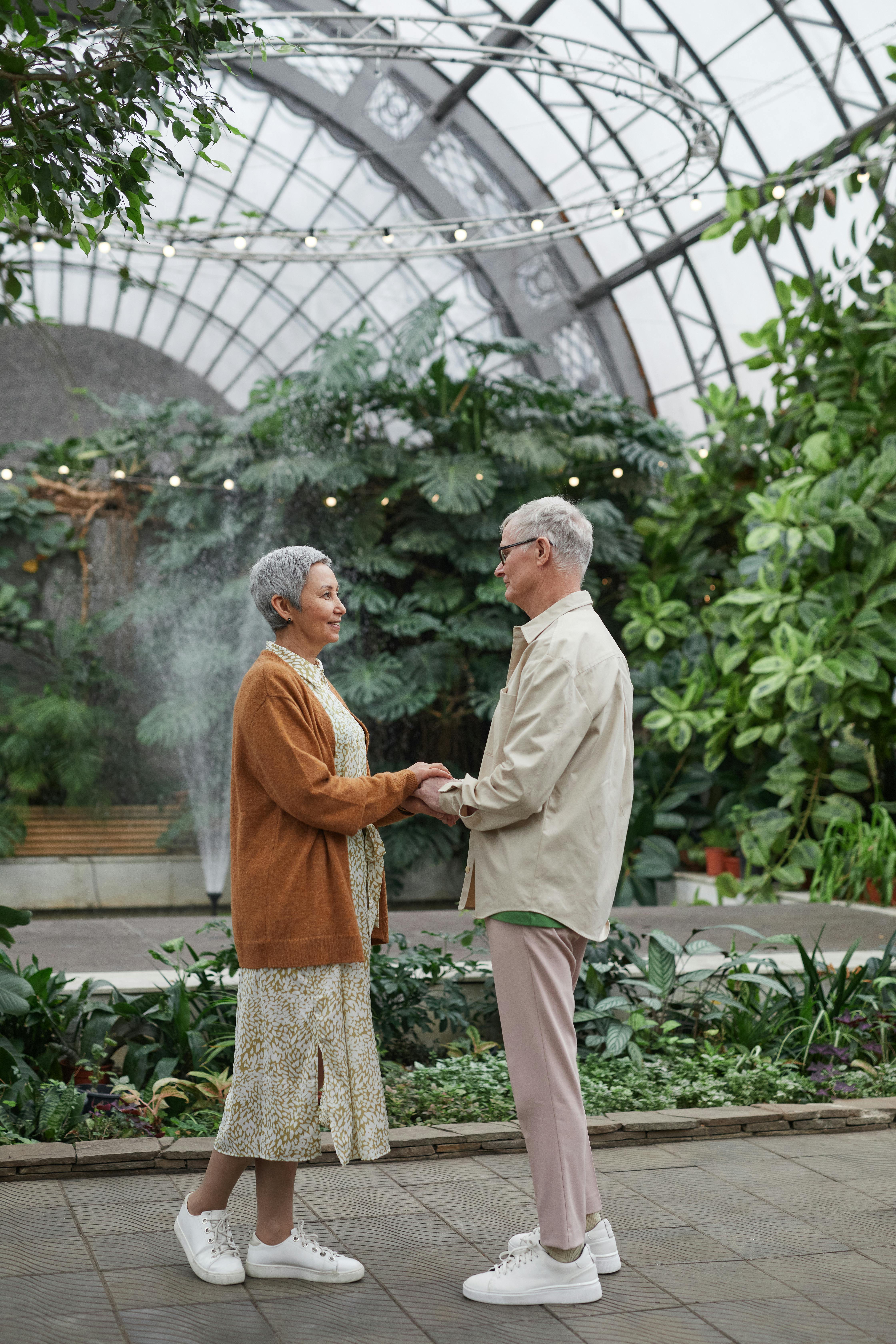 Senior couple stands in a lush greenhouse holding hands, symbolizing love and companionship.