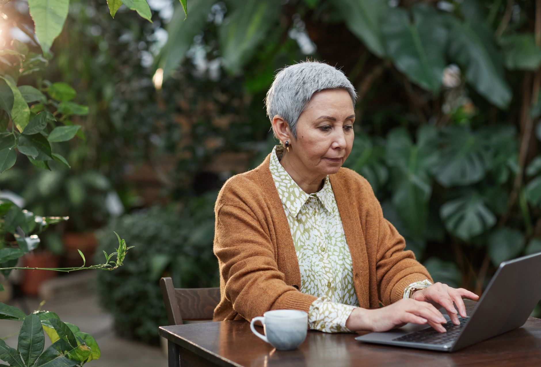 An elderly woman using a laptop outdoors surrounded by lush green plants.