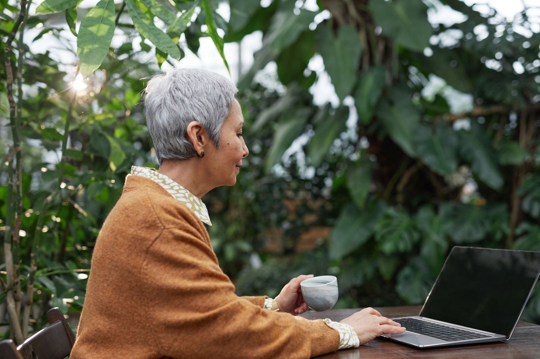 Senior woman with short silver hair using a laptop in a lush garden setting, holding a coffee cup.