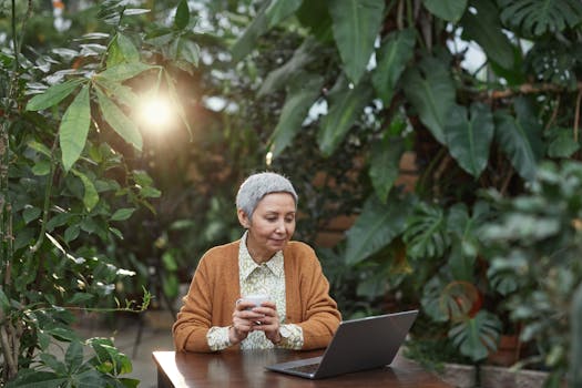 Senior woman enjoys coffee and laptop in a tropical garden, embracing nature and technology.